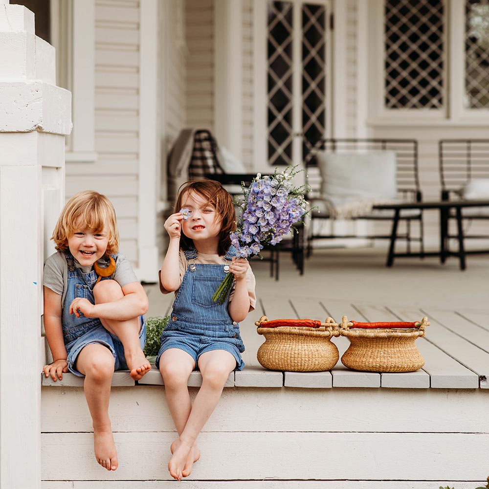 Bella Luna Toys Natural Bolga Basket sitting outside on a beige porch next to two kids in jean overalls holding purple flowers.